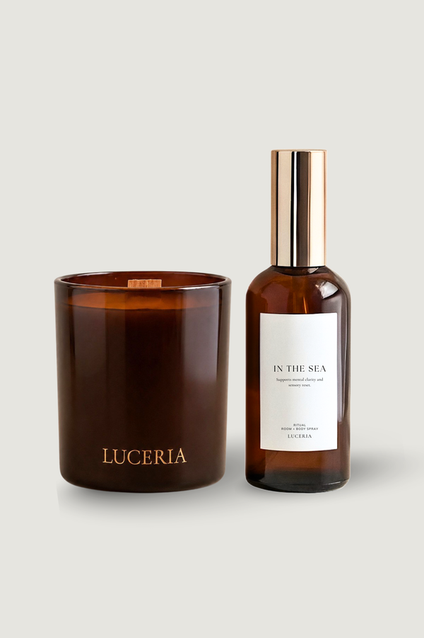 A dark amber glass candle jar and a matching amber bottle with a metallic gold cap and white label, both branded LUCERIA, on a light gray background.
