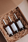 Three bottles of 'Repose', 'Woven', and 'Santal' in a cardboard box with shredded paper by Luceria Candle Co.