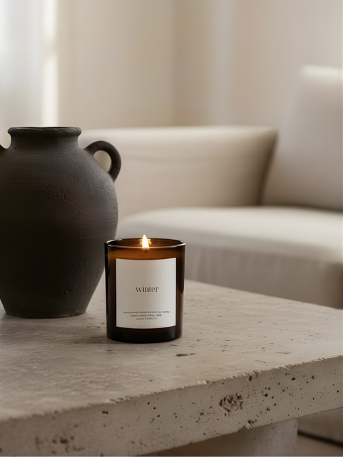 Candle with a label on a stone surface next to a black vase, with a neutral background
