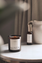 Amber candle labeled 'Woven' by Luceria on marble table with spray bottle and textured vase.