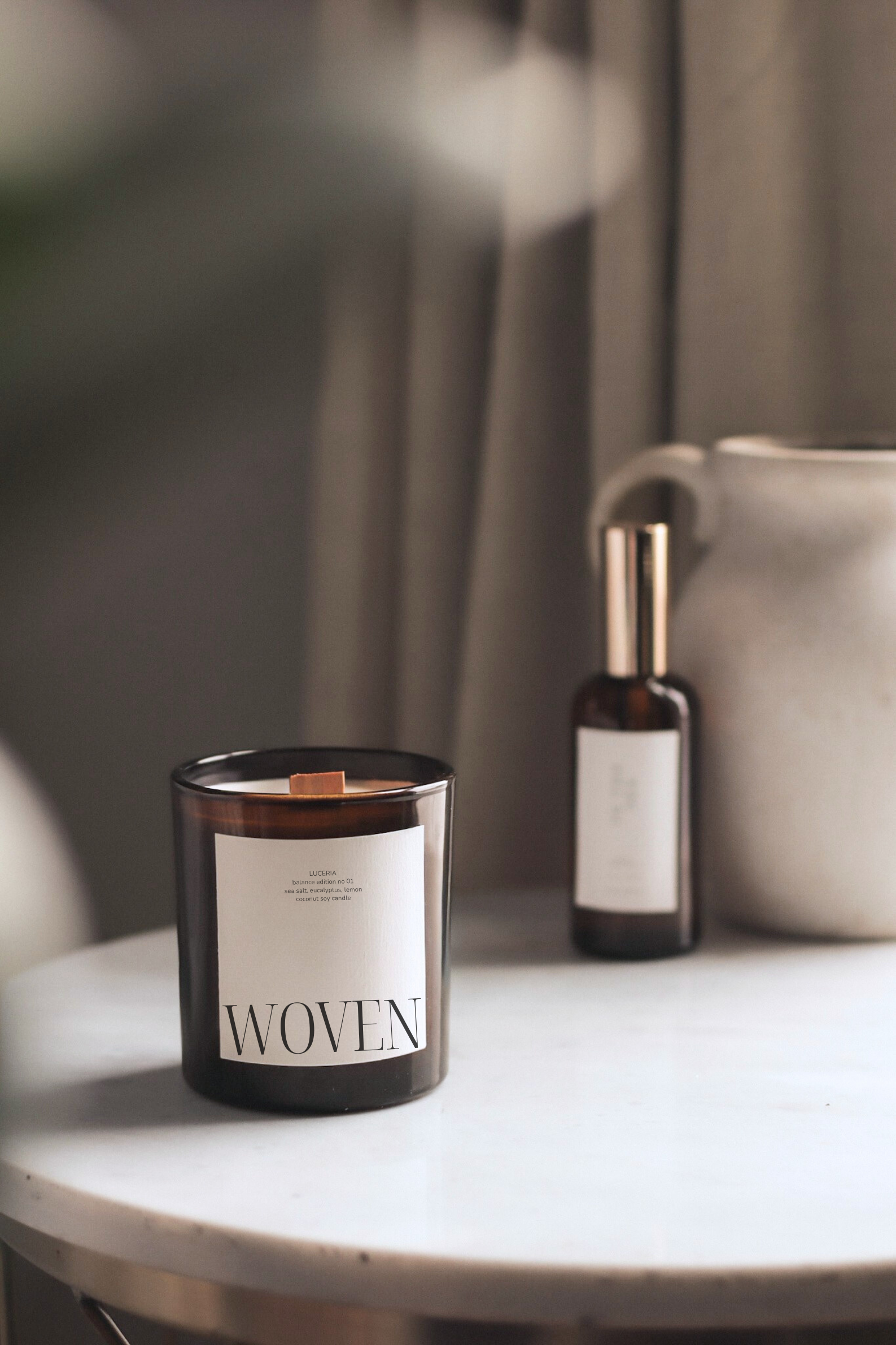 Amber candle labeled 'Woven' by Luceria on marble table with spray bottle and textured vase.